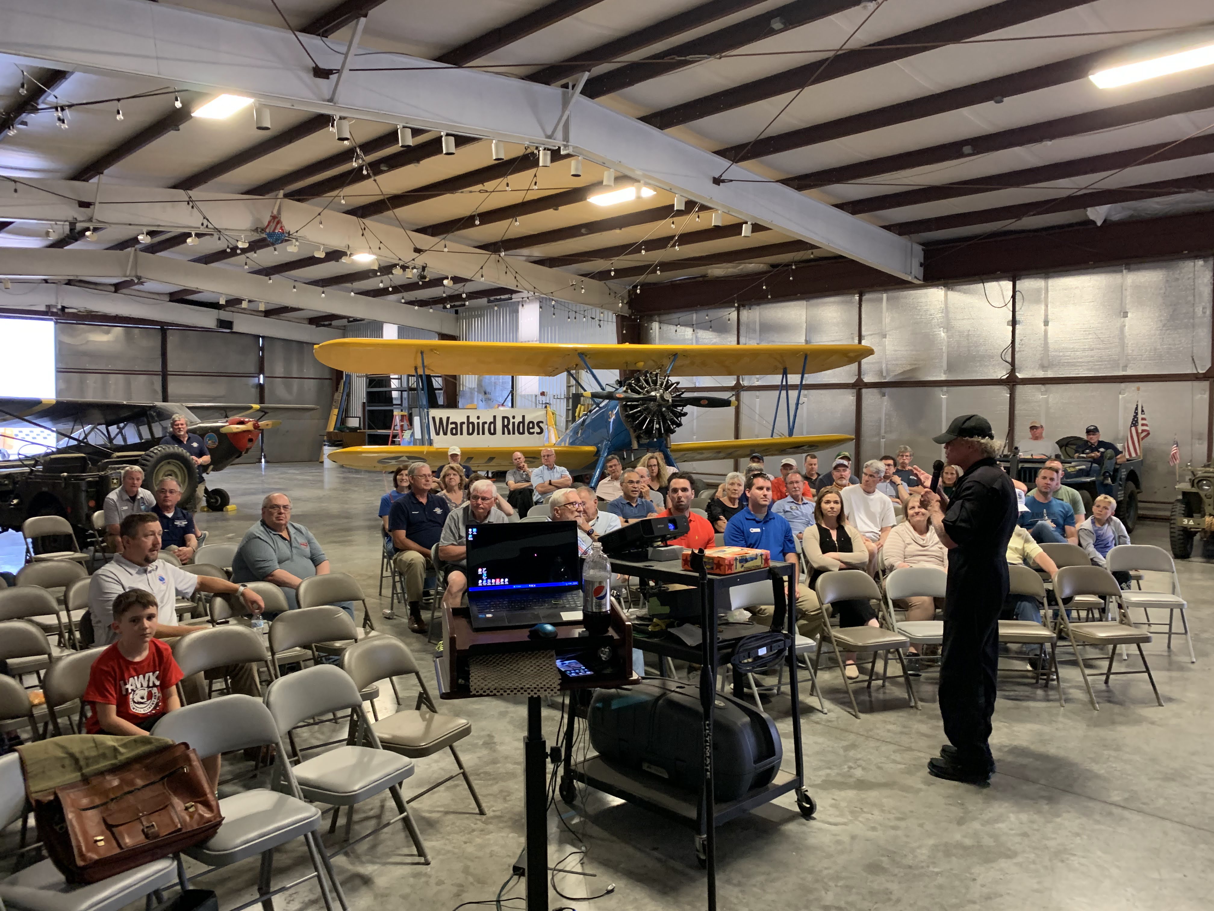 A person speaking to a group of people in a room with a plane.